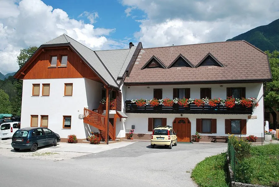 Bela hiša z dolgim balkonom in rdečim cvetjem, pred njo parkirišče, za njo zelen hrib